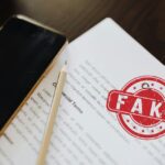 Documento contrattuale con timbro rosso "FAKE", smartphone e parte di laptop. Immagine che simboleggia polizze fideiussorie false e truffe digitali segnalate dall'IVASS.